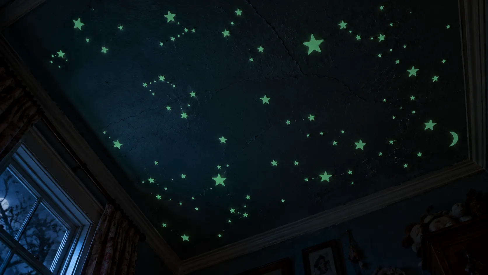 Lying on the floor looking up at a bedroom ceiling covered in glow-in-the-dark stars, a moon, a window with a tree outside.